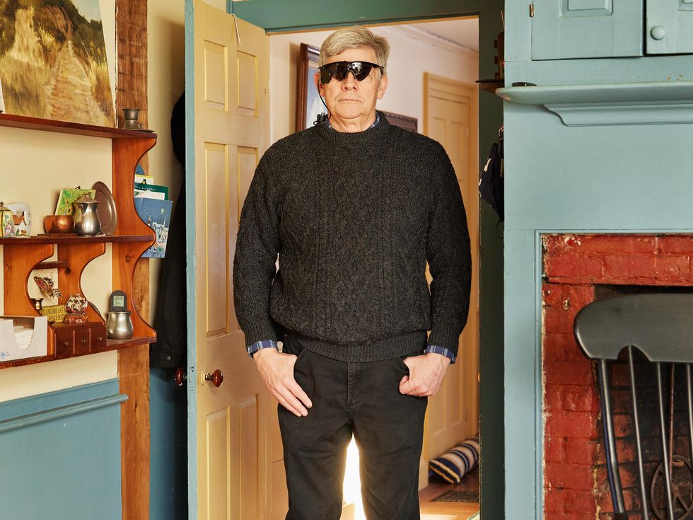 A man wearing bulky black sunglasses stands inside a living room.