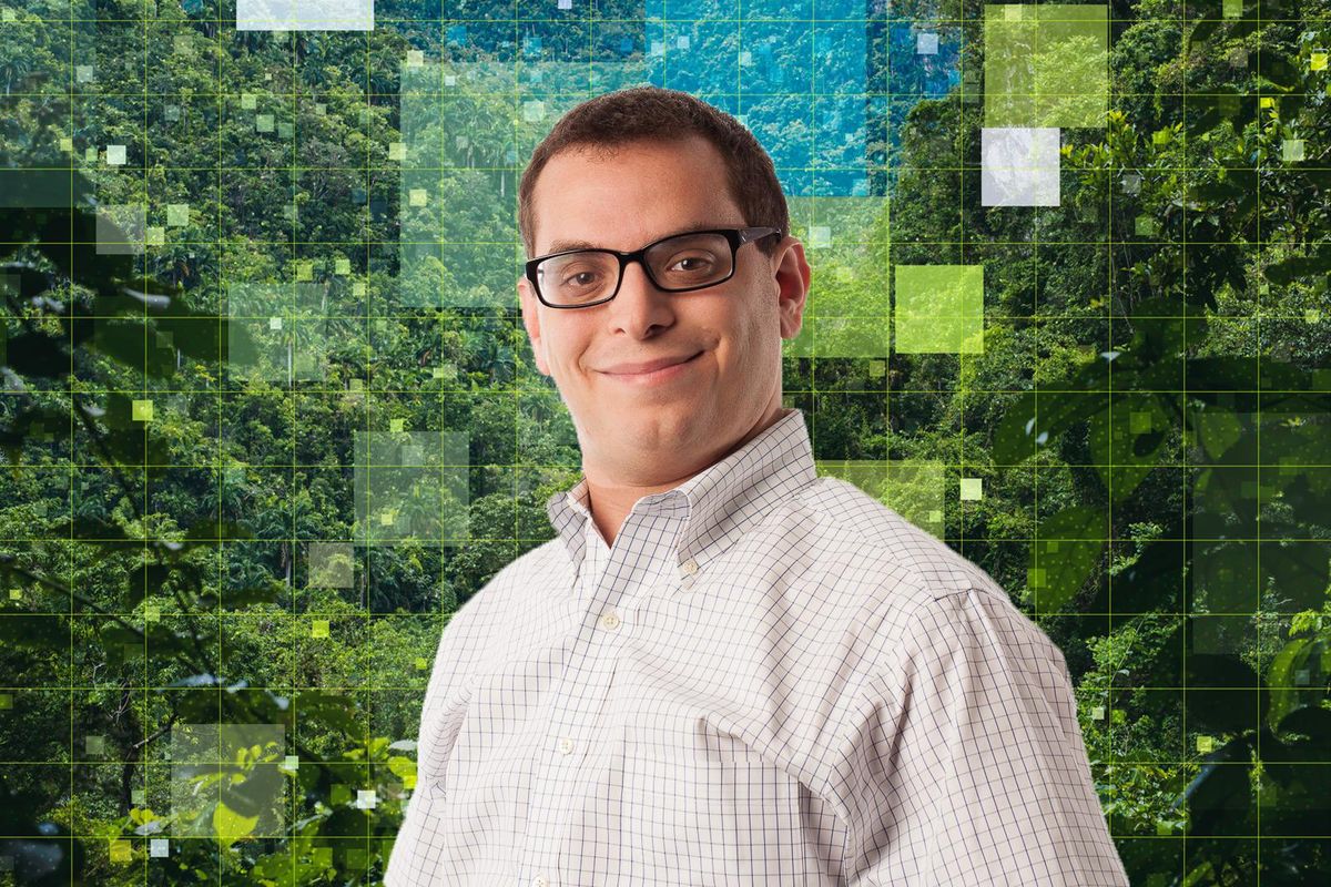 A man with glasses and a checkered shirt smiles in front of a pixelated background