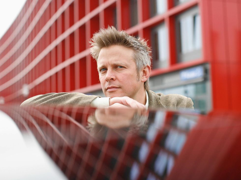 A man with spiky blond hair stands in front of a red building which is also reflected on a surface he is leaning against.