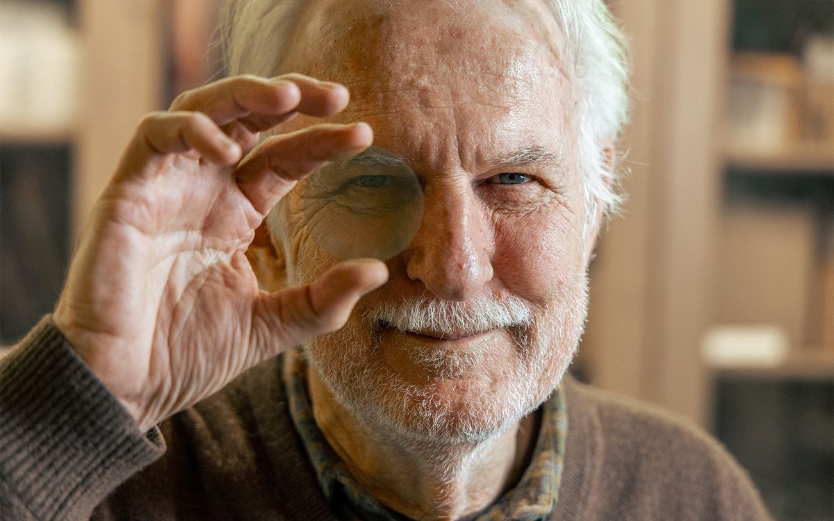 A man with white hair, beard and mustache holds a circular piece of material in front of his eye.