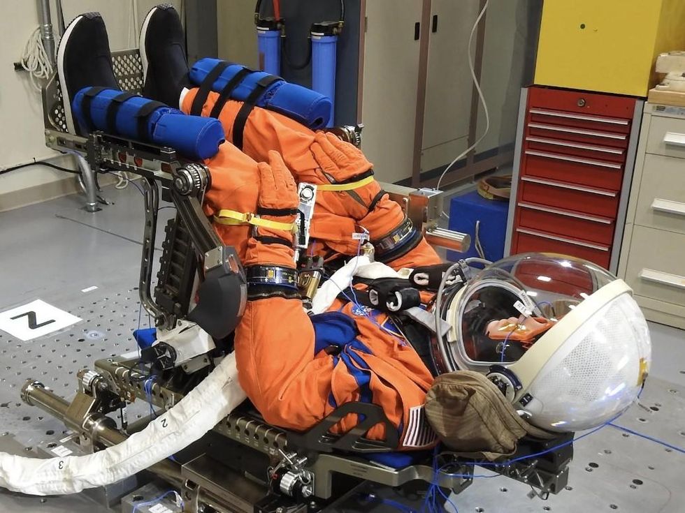 A mannequin in an orange flight suit lies on its back in a testing room
