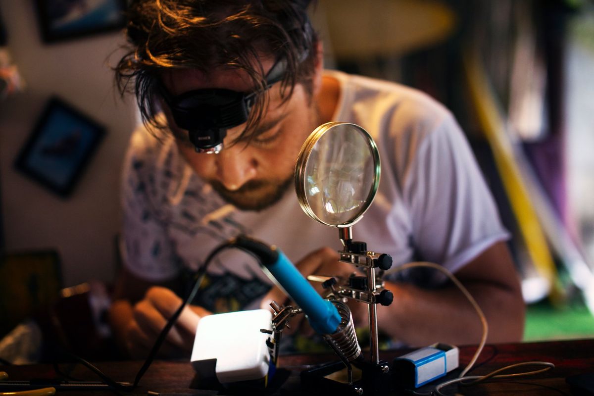 A mean wearing a headlamp inspects an electronic device on a desk that includes a magnifying glass and a soldering iron.