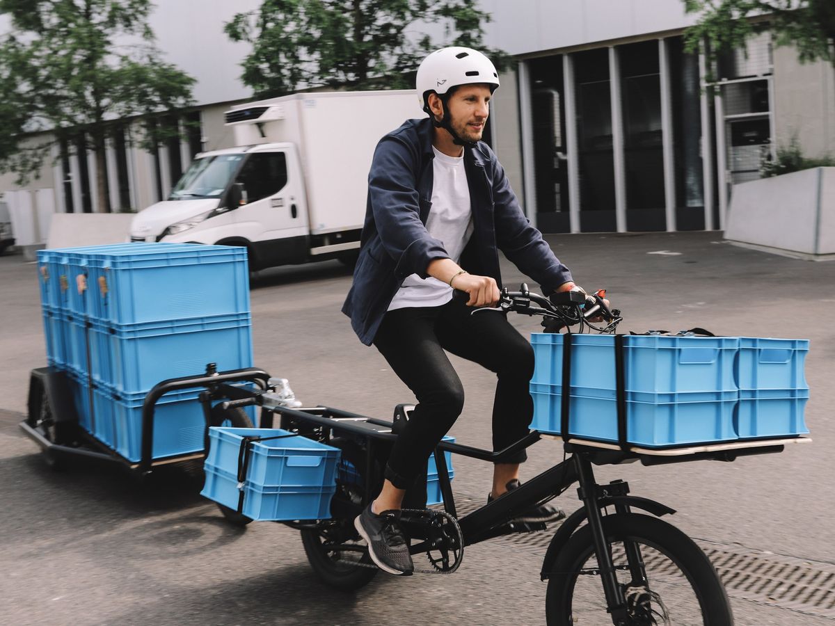 a mean wearing a white bike helmet pedaling a bike with blue containers on front, the sides and behind in a trailer in a street scene