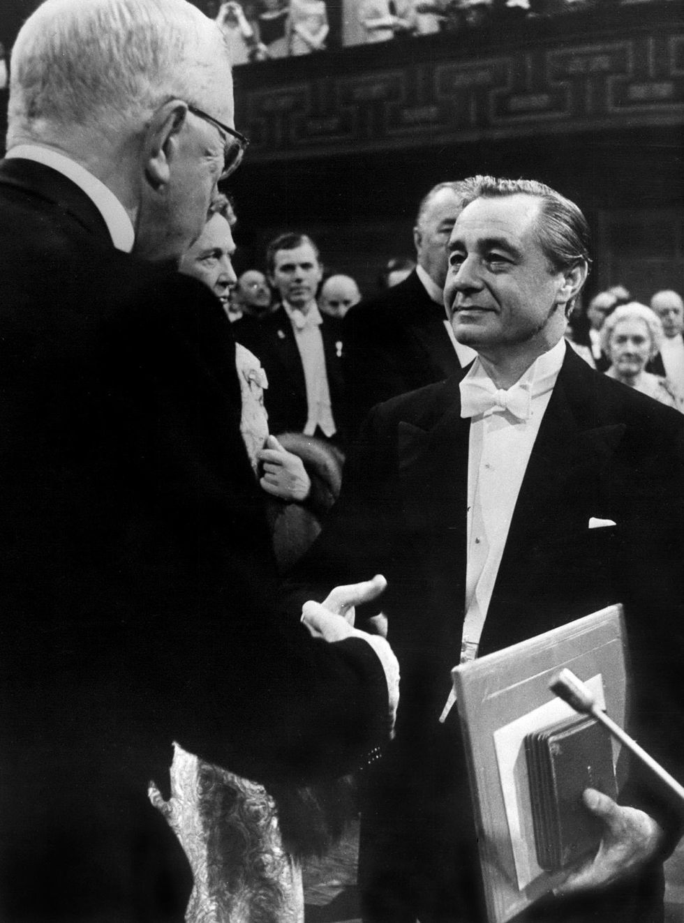 A middle-aged man in a tuxedo shakes hands with a white-haired man while a well-dressed crowd looks on.