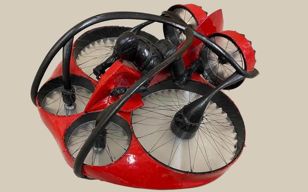 A model of a circular red craft with six rotors arranged around its edge and a black-suited person straddling the craft's middle.