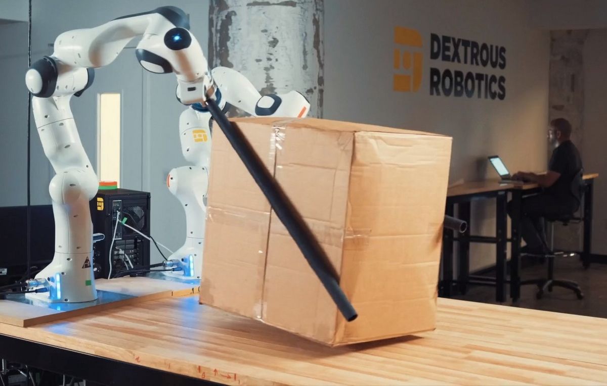 A pair of white robot hands each have a black tube attached. The tubes are pressed against the sides of a cardboard box to lift it.
