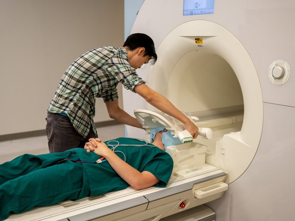 A person in a plaid shirt fits a helmet apparatus on top of the head of another person who is lying at the entrance of an MRI machine.