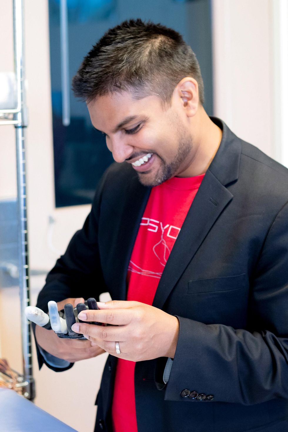 A person in a red shirt and jacket smiling and looking at a prosthetic hand.