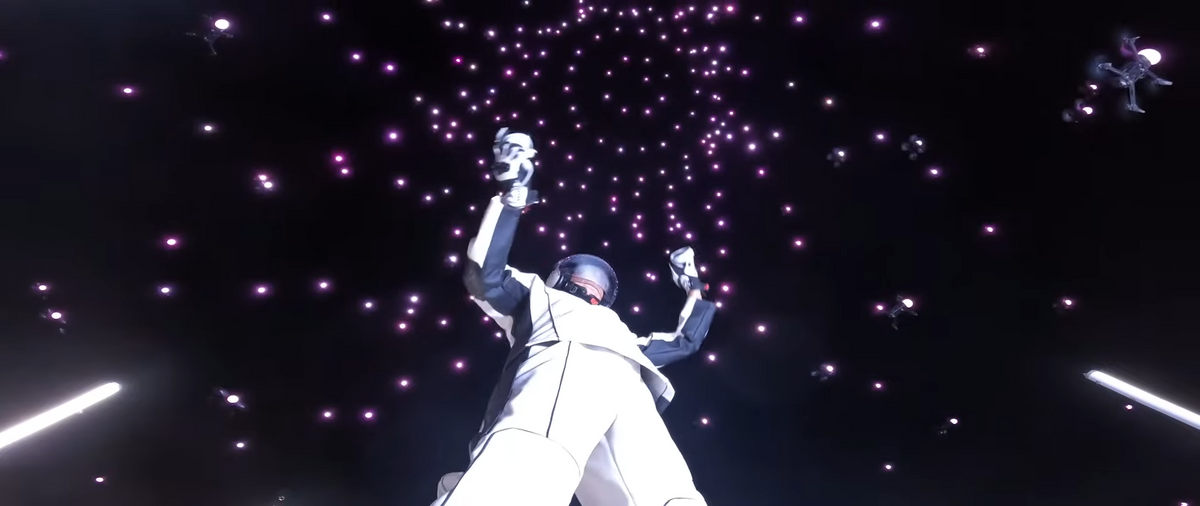 A photo from a low angle looking upwards of a human in a futuristic suit gesturing at night while hundreds of illuminated drones swarm above him