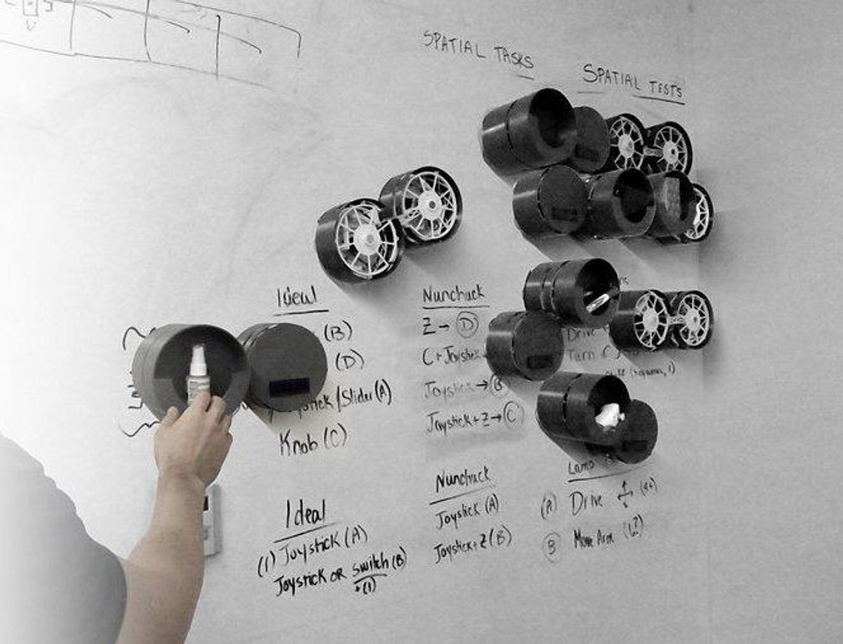 A photo of a dozen small round robots with containers for items attached to a whiteboard