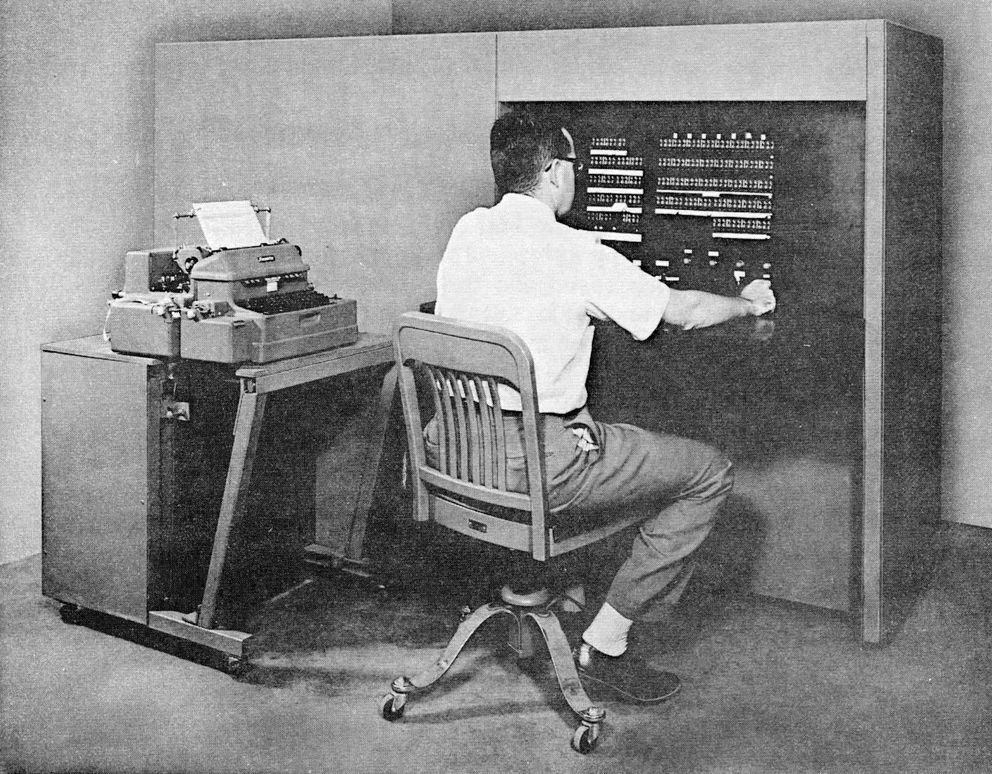 A photo of a man sitting at an early computer next to a typewriter.