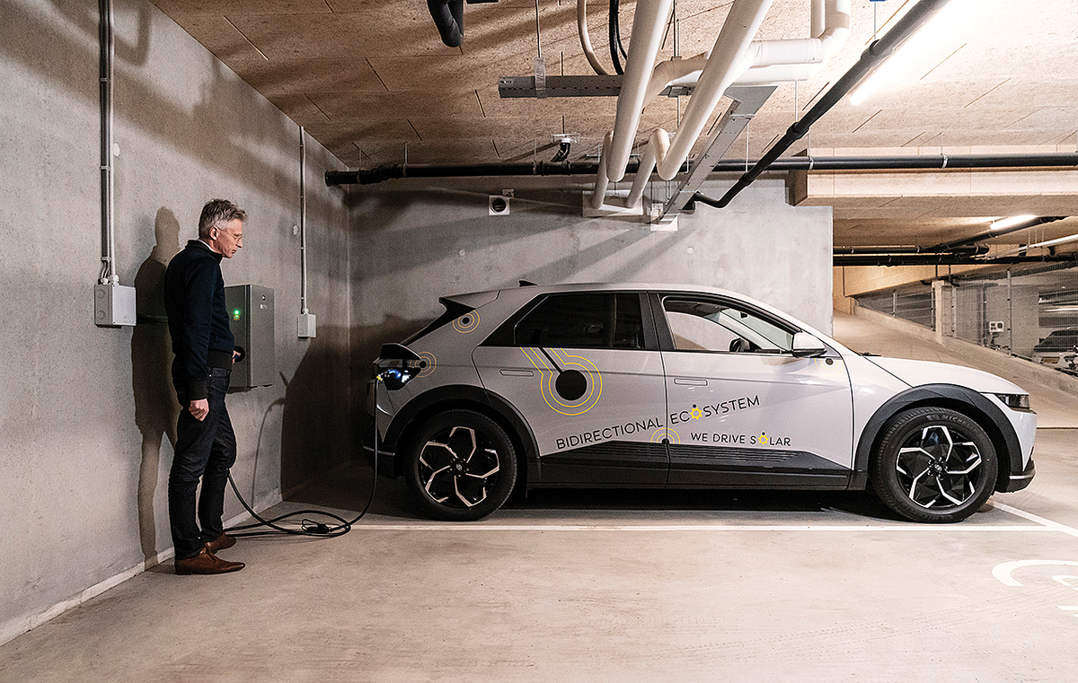 A photo of a man standing next to a charging station near an electric car that has "Bidirectional Ecosystem" on this side.