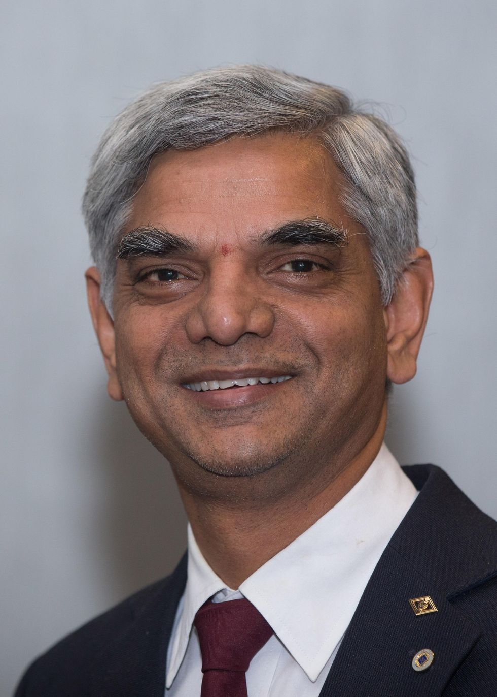 A photo of a man with grey hair and a blue jacket and red tie.