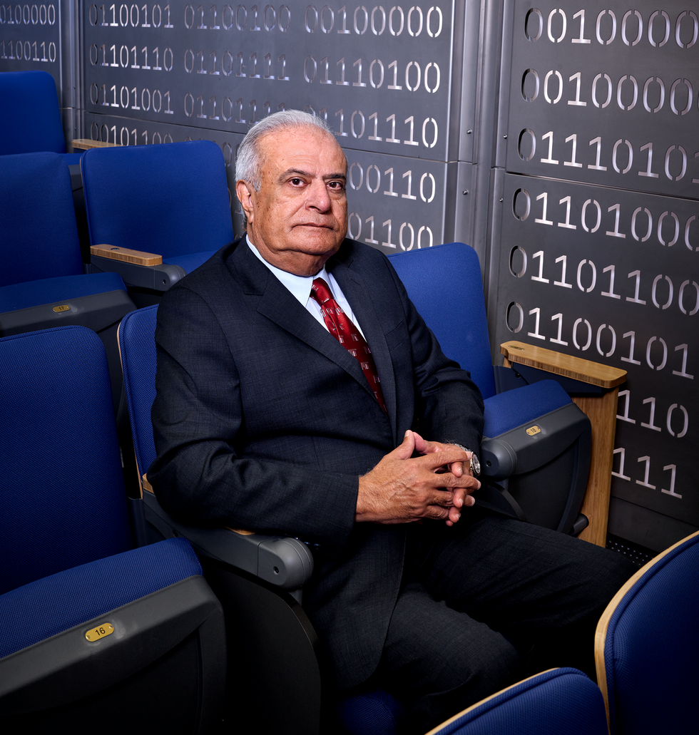 A photo of a seated man in a dark suit with binary numbers on the wall behind him.