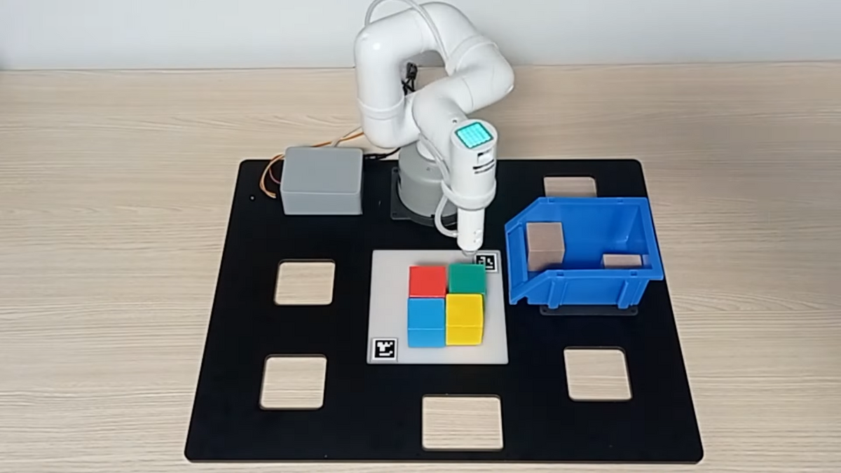 A photo of a small white robot arm on a desk having assembled the Microsoft logo out of colored blocks