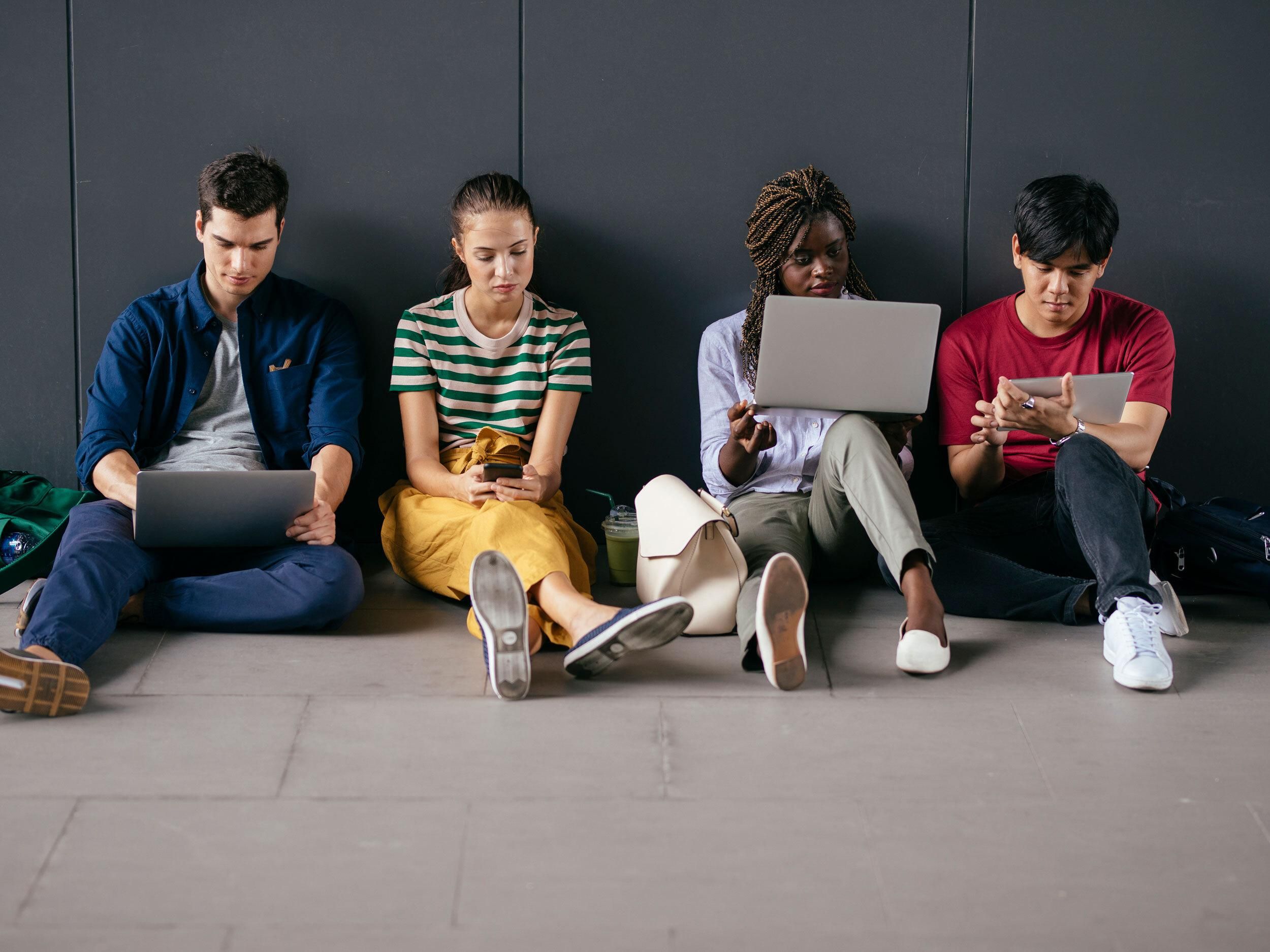 A photo of four people sitting against a wall with a variety of different devices in their hands.   