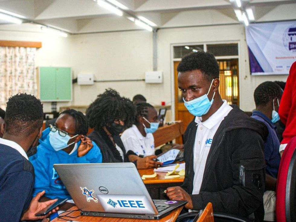A photo of people in a classroom and a person in an IEEE shirt and mask looking at a laptop with an IEEE sticker on it.  