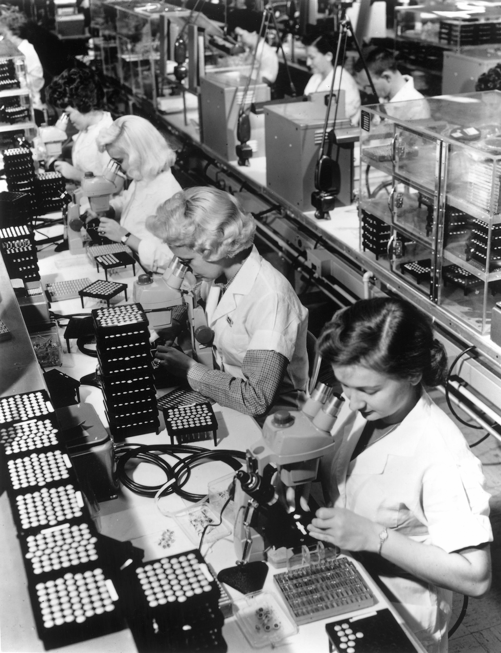 A photo of rows of people sitting in front of microscopes and stacks of transistors.
