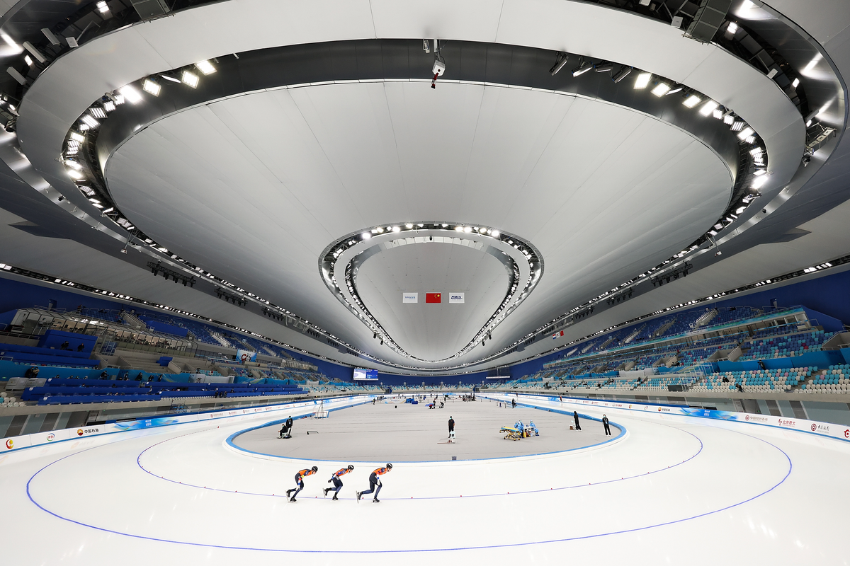 A photo of the interior of a large ice skating rink, with three skaters in the foreground