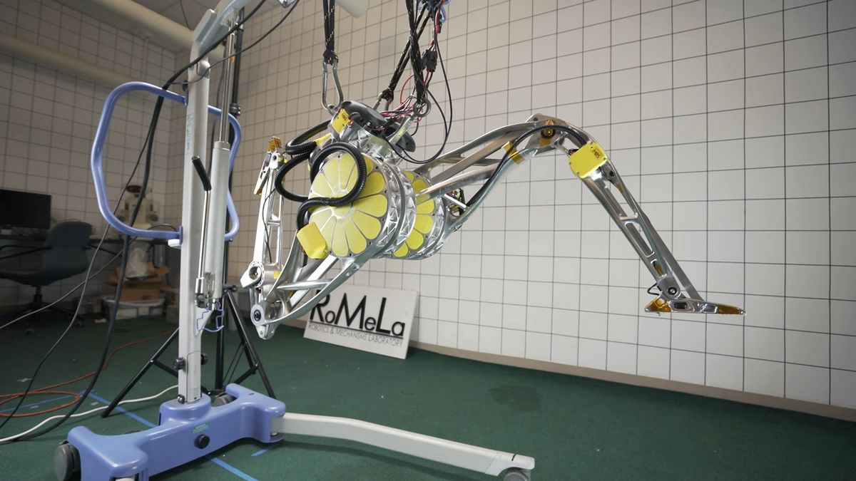A photo of the lower torso and legs of a humanoid robot in a dynamic running position suspended on a test stand in a lab