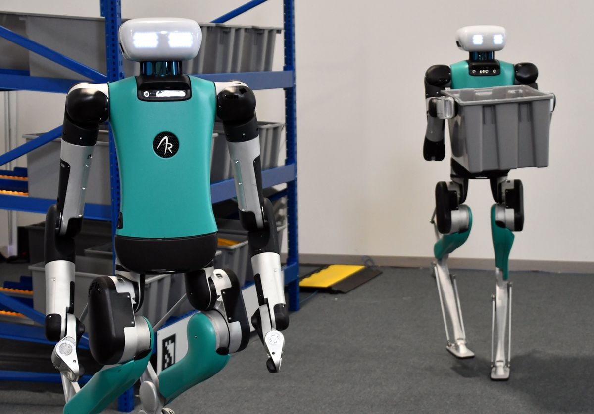 A photo of two bipedal robots moving grey plastic bins around a model warehouse