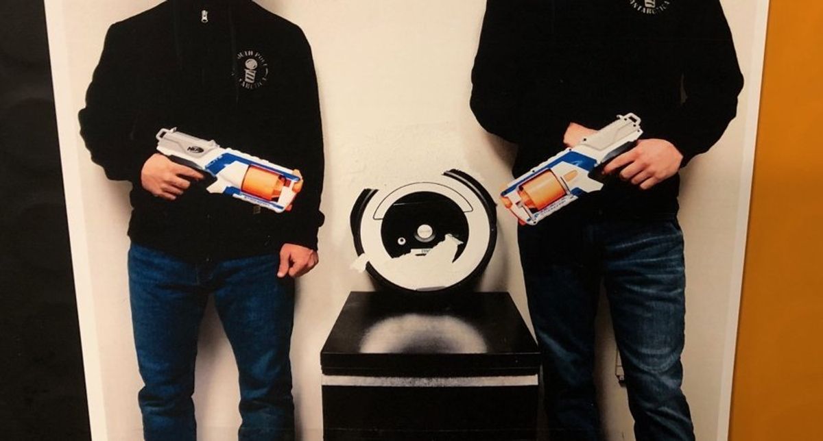A photo of two men from the shoulders down standing and pointing Nerf guns at a Roomba on a pedestal between them.