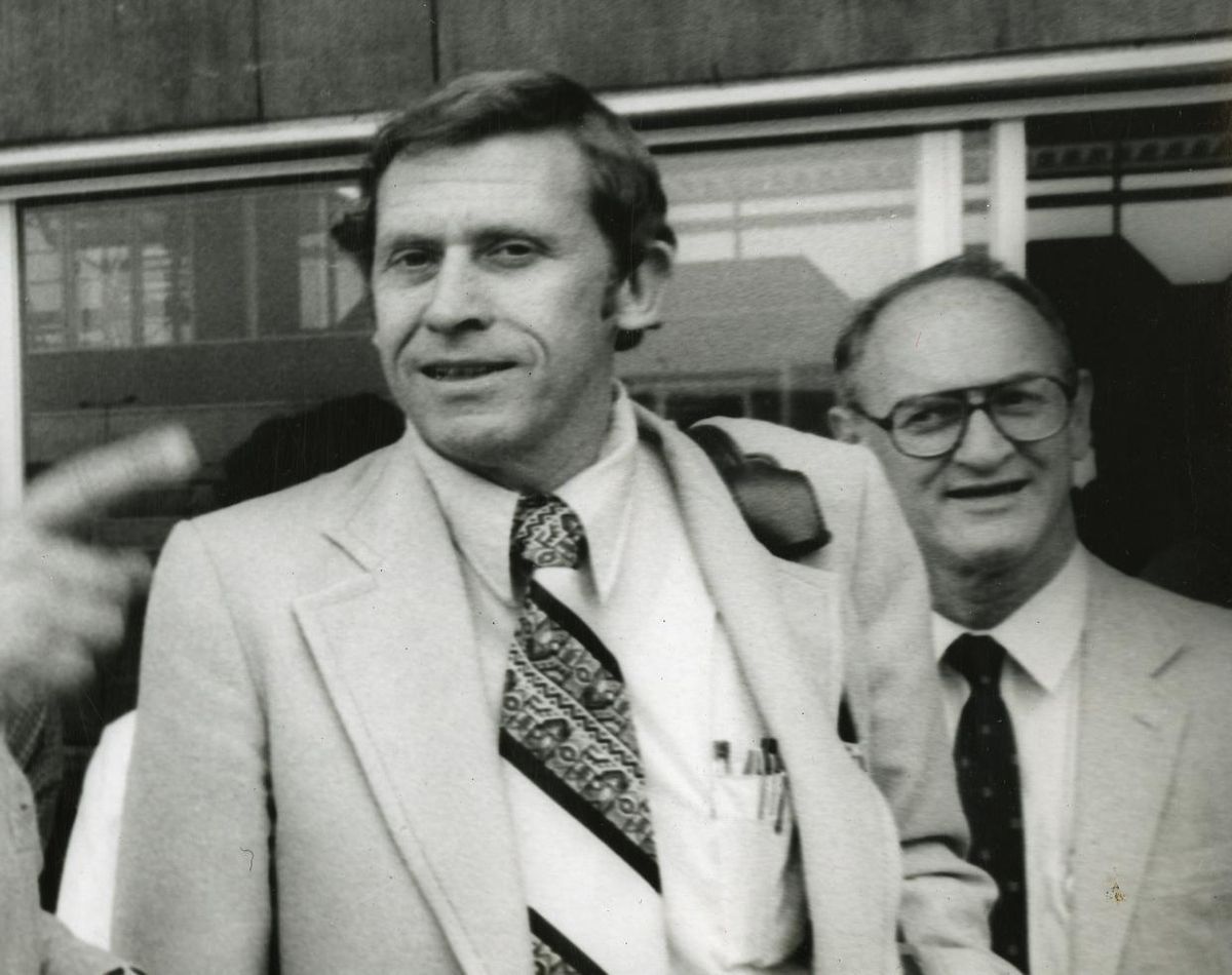 A photo of two men in suits.  One behind the other.  