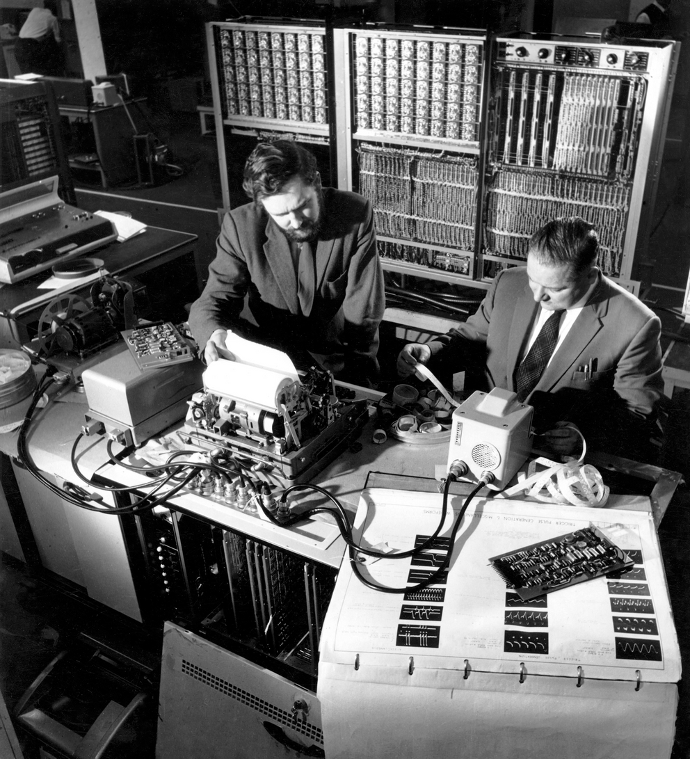 A photo of two men sitting at a terminal in front of an early computer.