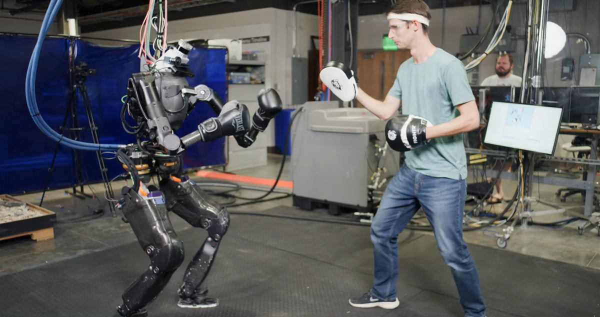 A photo showing a humanoid robot wearing boxing gloves facing a human wearing hand pads.