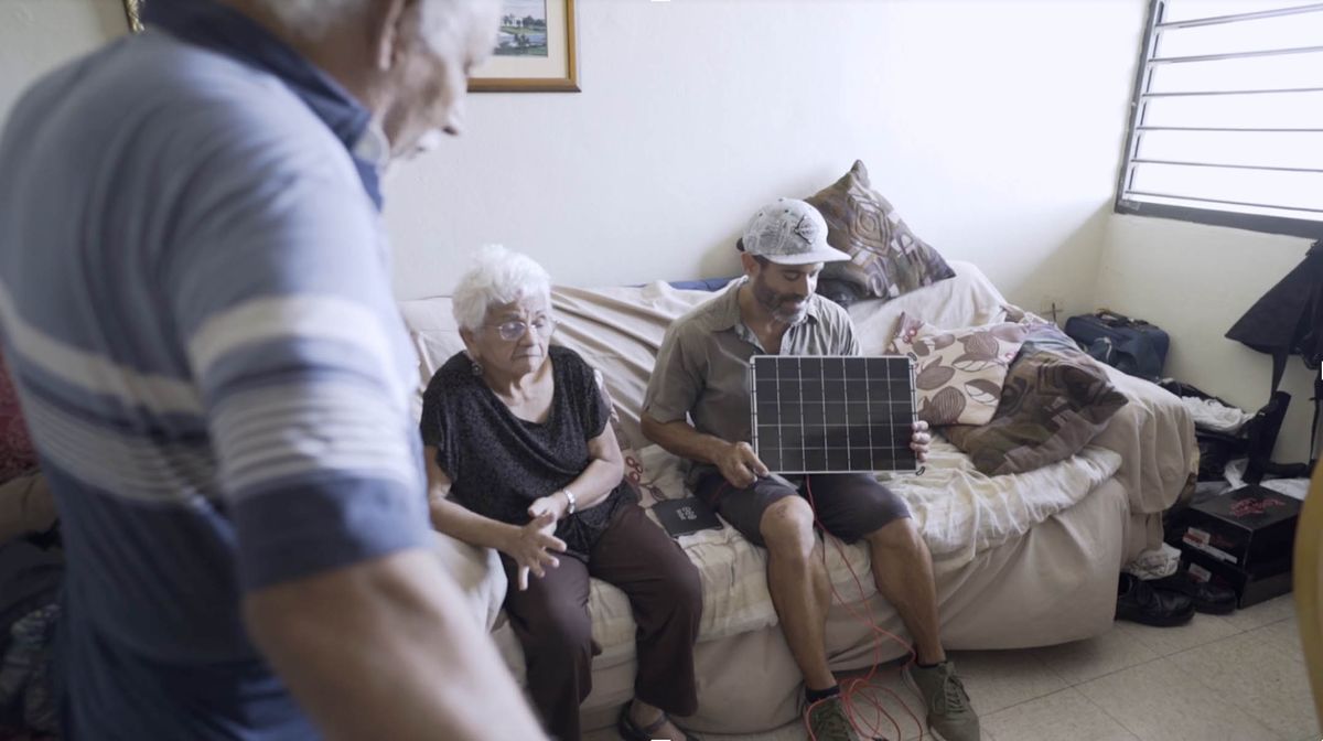 A photo shows a man sitting on a couch holding a solar charger in front of residents at a senior center.