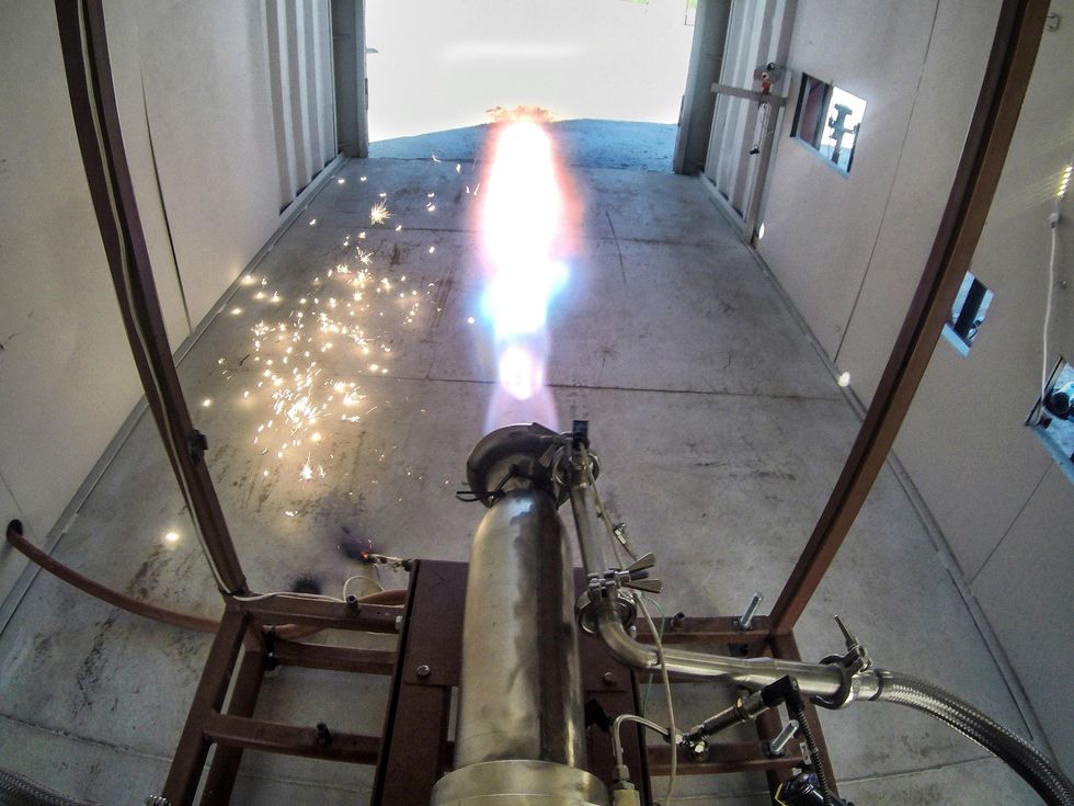 A photo shows a metal engine nozzle with a jet of fire coming out of one end. 