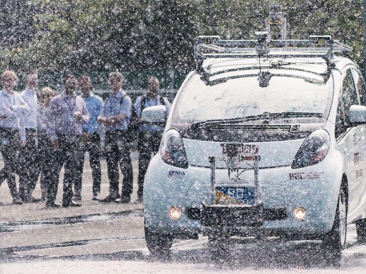 A photo shows a white self-driving car obscured by rain coming from nozzles above it, with people standing in the background to watch.