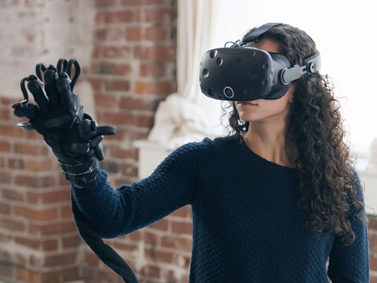 A photo shows a woman wearing the new HaptX glove, which resembles a large black ski glove with wires attached to it, and an HTC Vive headset. 