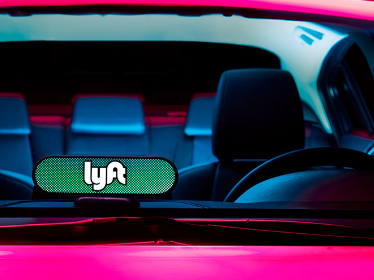 A photo shows the black interior of a hot pink car, and a sign with the Lyft logo mounted to the dashboard. 