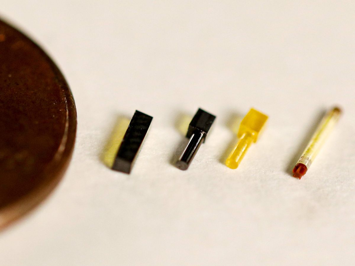 A photograph of several tiny shapes. From left, they are half of a brown disc, a black rectangular prism, a black rectangular prism with a cylinder on one end, a yellow rectangular prism with a cylinder on one end, and a clear cylinder with red end caps.