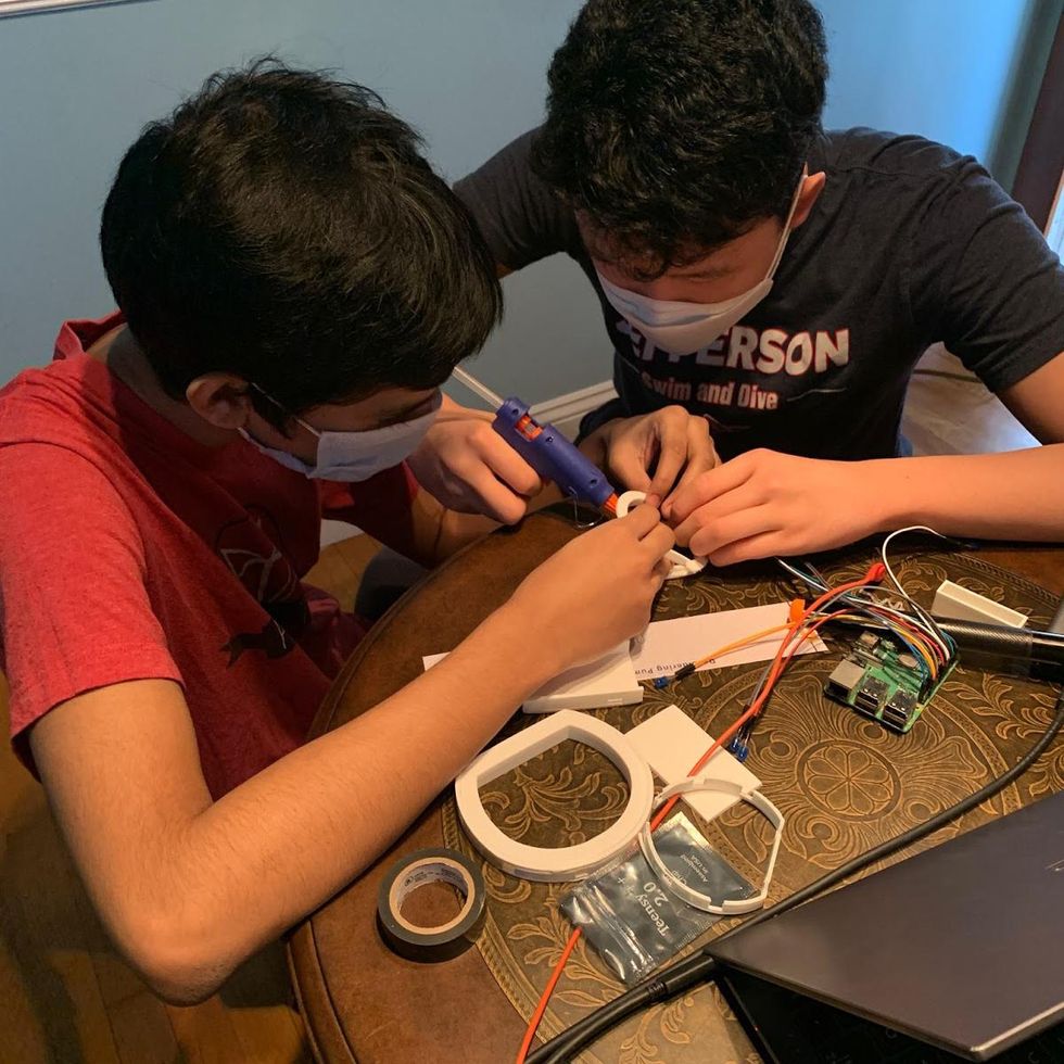 A photograph of two young males wearing surgical masks sitting at a desk gluing together parts with various other electronic parts placed on the surface. Placement: Anywhere needed in later sections