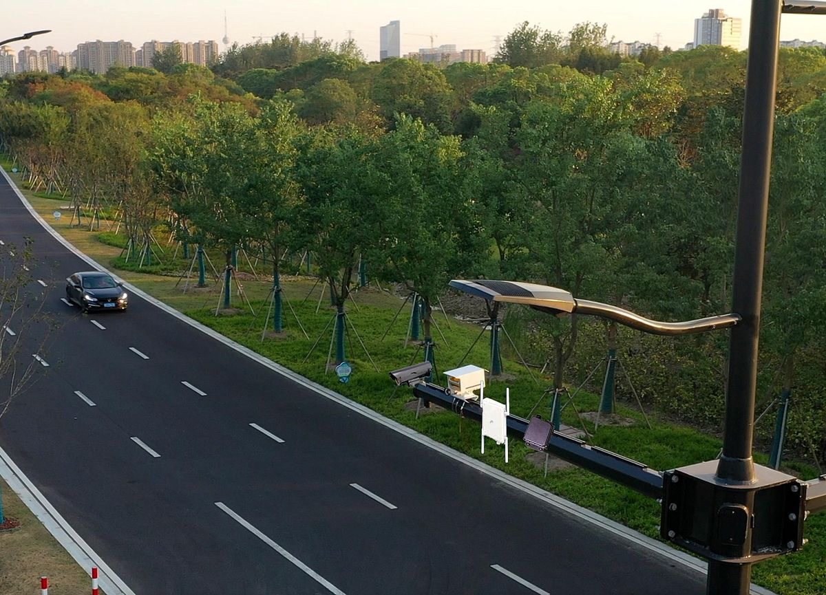 A photograph shows a single car headed toward the viewer on the rightmost lane of a three-lane road that is bounded by grassy parkways, one side of which is planted with trees. In the foreground a black vertical pole is topped by a crossbeam bearing various instruments. 