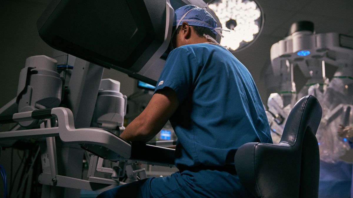 A physician positions his head into a special medical display and uses his hands to remote operate a surgical robot, seen in the background of an operating room.