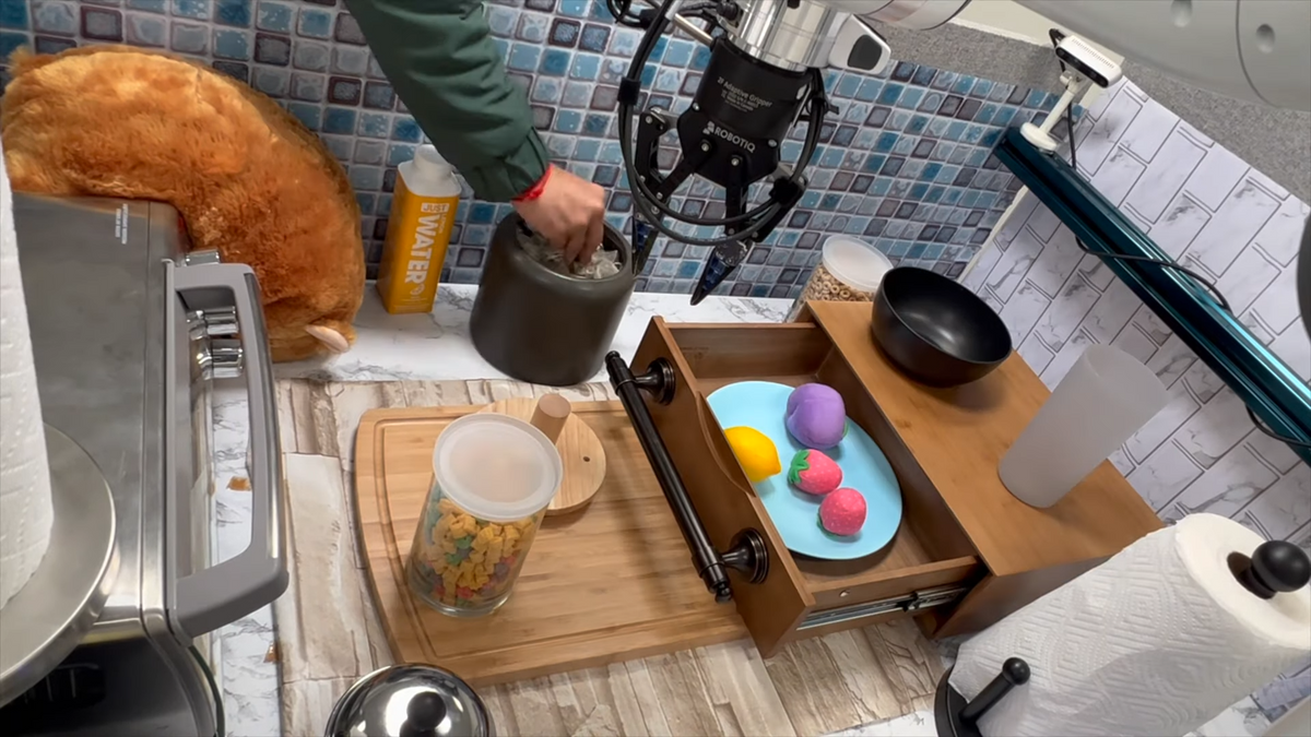 A picture of a robot arm and a human arm collaborating in a fake kitchen that includes a drawer, toaster oven, cutting board, and a jar of Cap'n Crunch