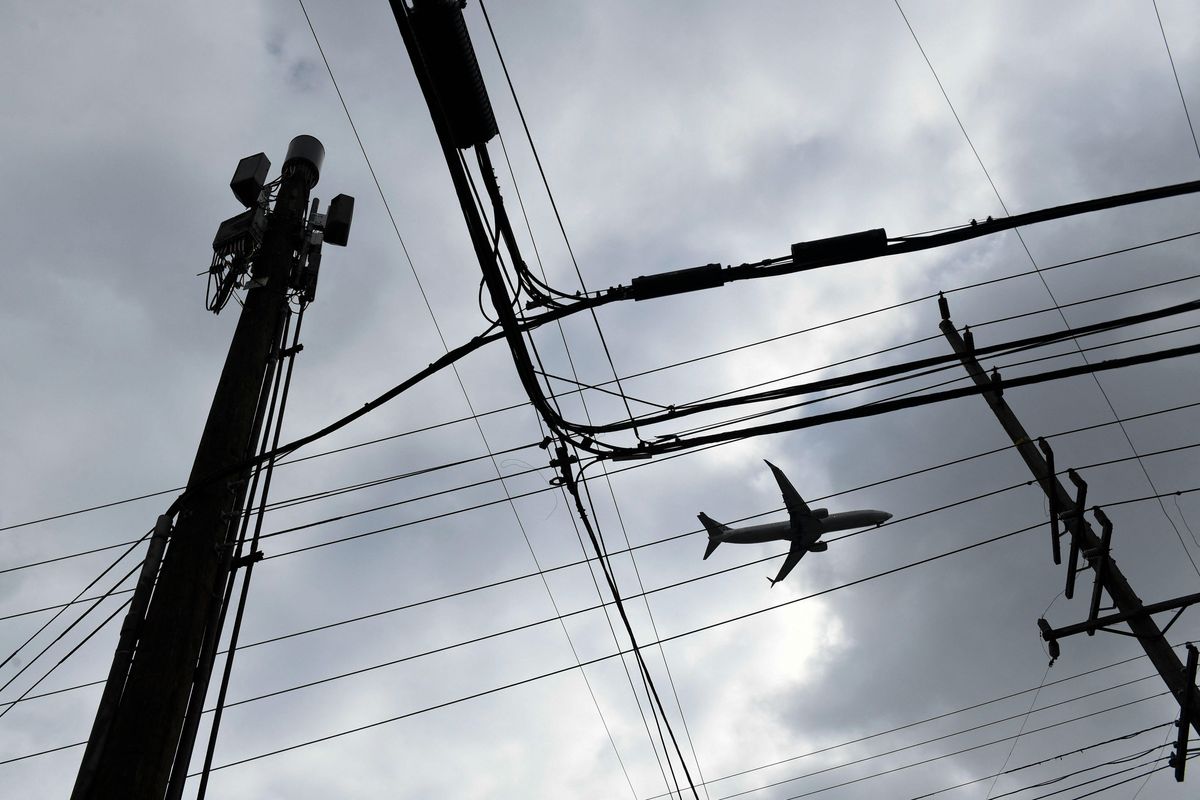 A plane, a 5G cell tower, and electrical wires are seen in silhouette against a cloudy sky