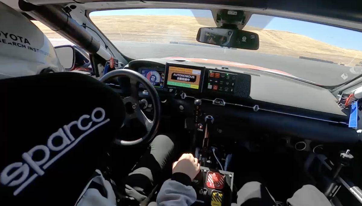 A professional driver wearing a helmet sits behind the steering wheel, without touching it, of a customized Toyota sports car that is drifting autonomously on a closed asphalt track.