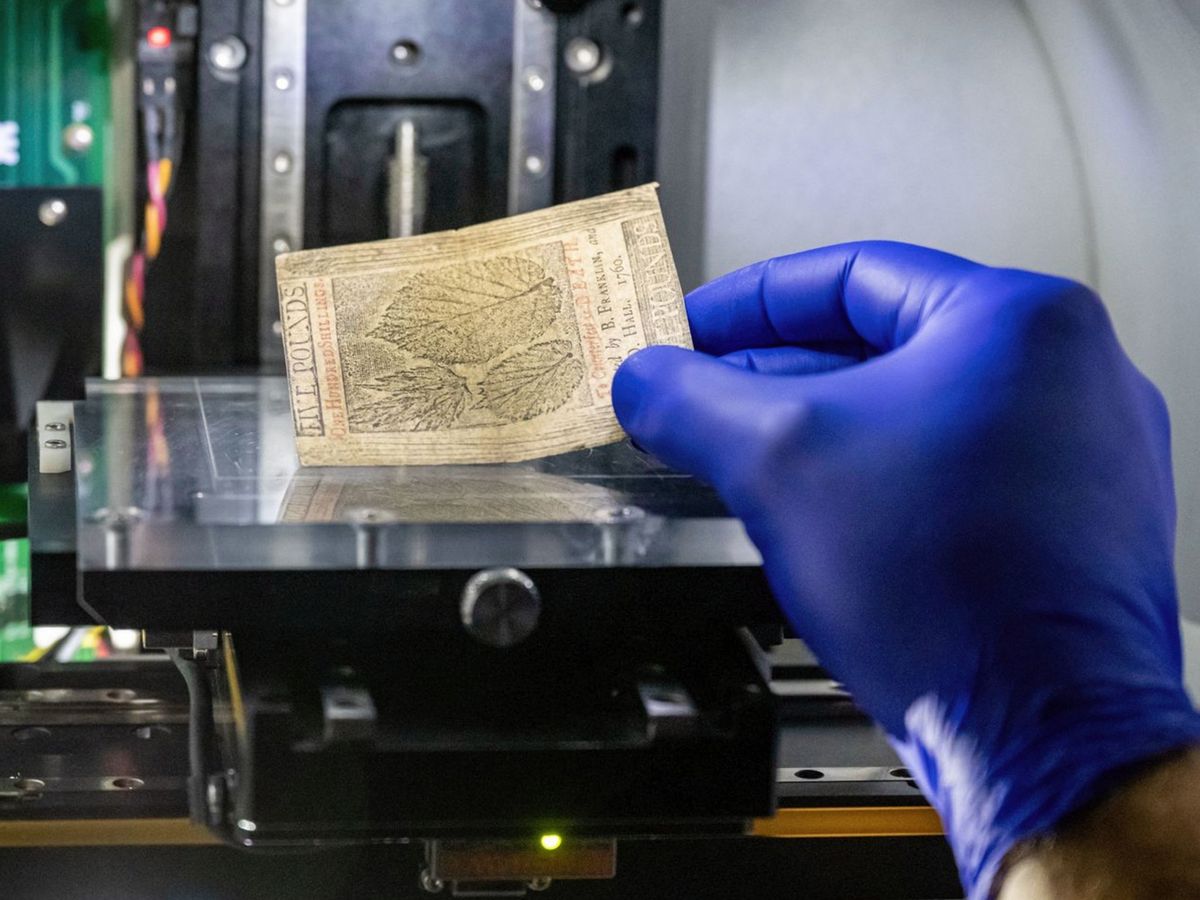 A purple gloved hand holds a rectangle of old paper currency on top of lab equipment.