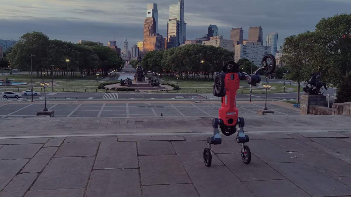 A red bipedal robot with wheels for feet and hands stands upright at the top of steps with the city of Philadelphia in the background
