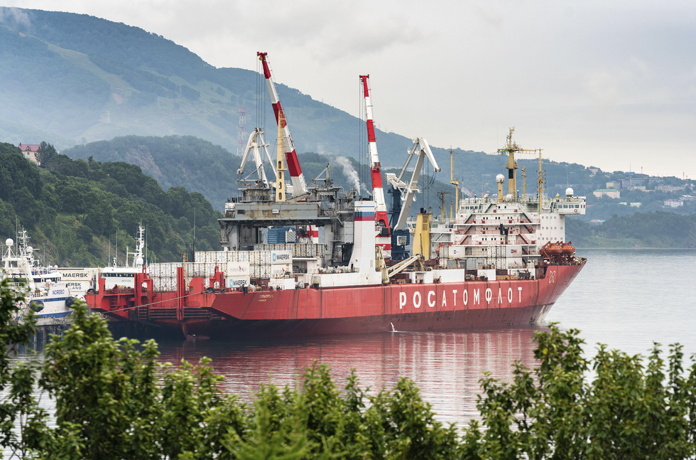 A red cargo ship is shown in a harbor surrounded by mountainous terrain.