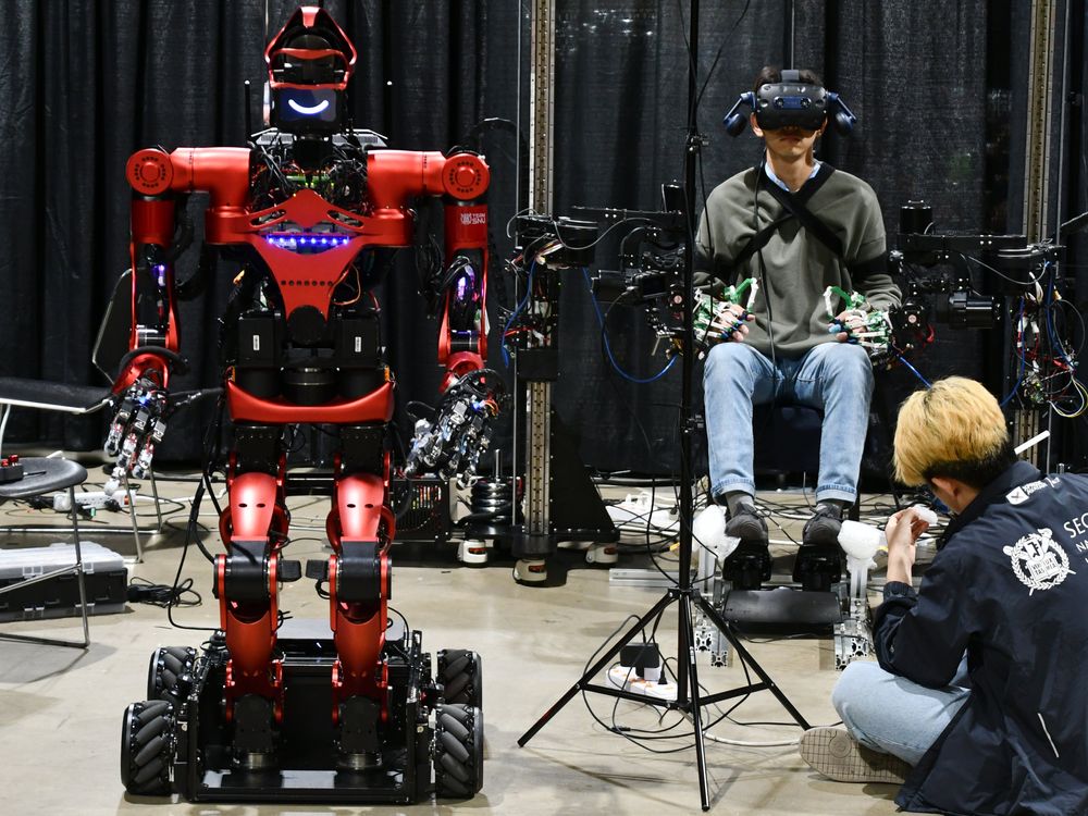 A red humanoid robot on a wheeled platform waits beside a human wearing a VR headset with his hands and arms covered in mechanical sensors.