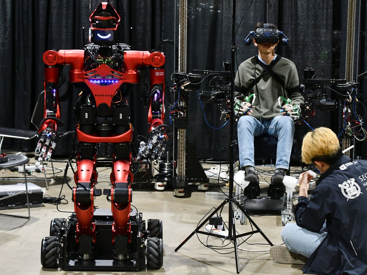A red humanoid robot on a wheeled platform waits beside a human wearing a VR headset with his hands and arms covered in mechanical sensors.