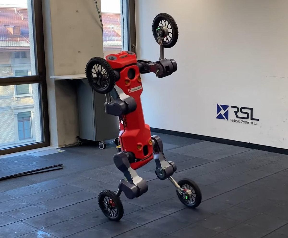 A red quadruped robot with wheels for feet balances upright on its hind legs in an empty room.