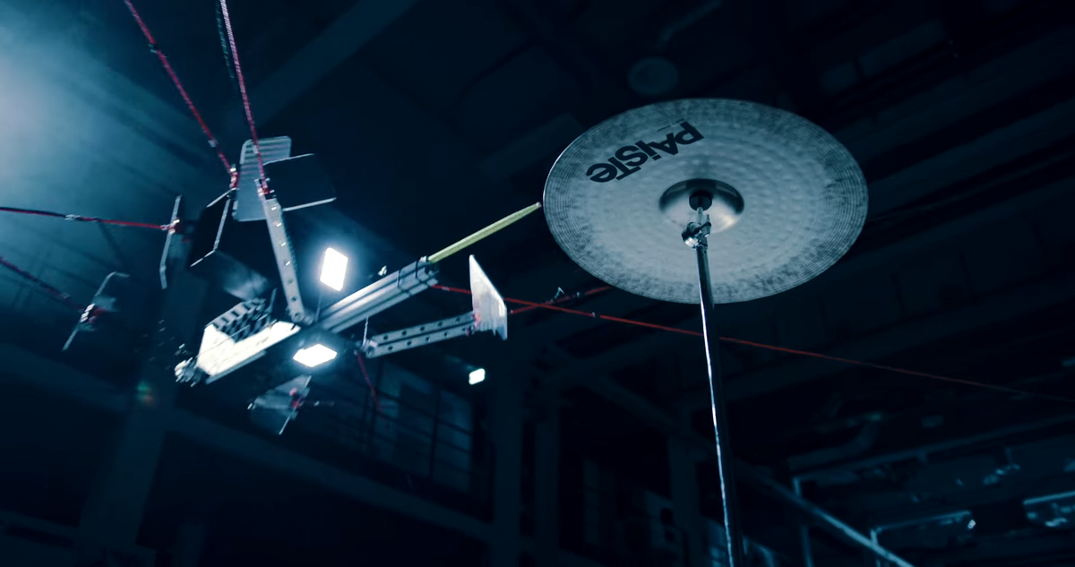A robot suspended by red cables holding a drumstick descends towards a cymbal in a dramatically lit warehouse
