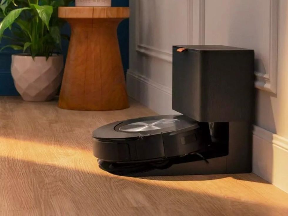 A Roomba sits on its charging base, which also empties the robot's dust bin, on a living room with wooden floors.