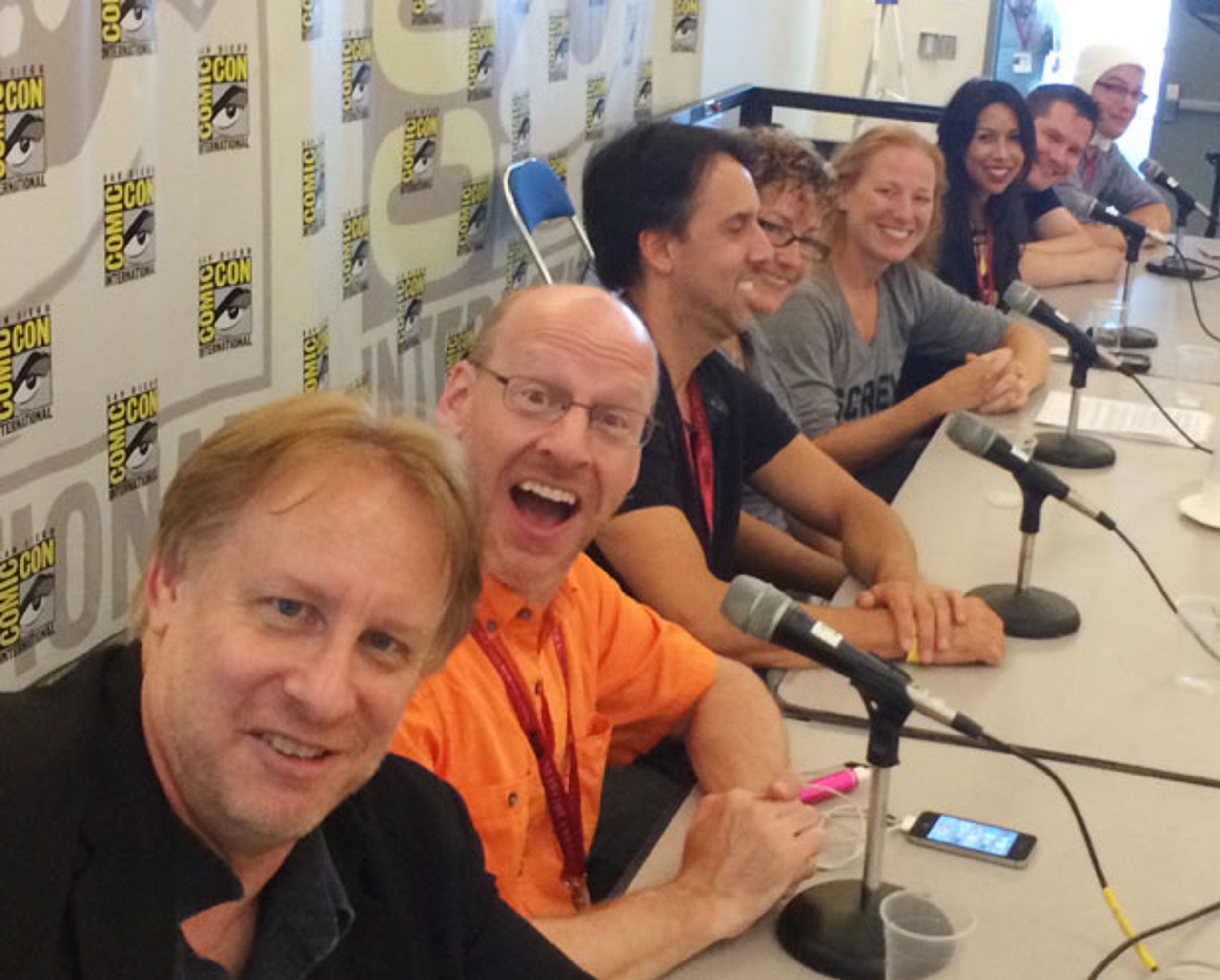 A row of 8 smiling people sitting at a table with a San Diego Comic backdrop behind them
